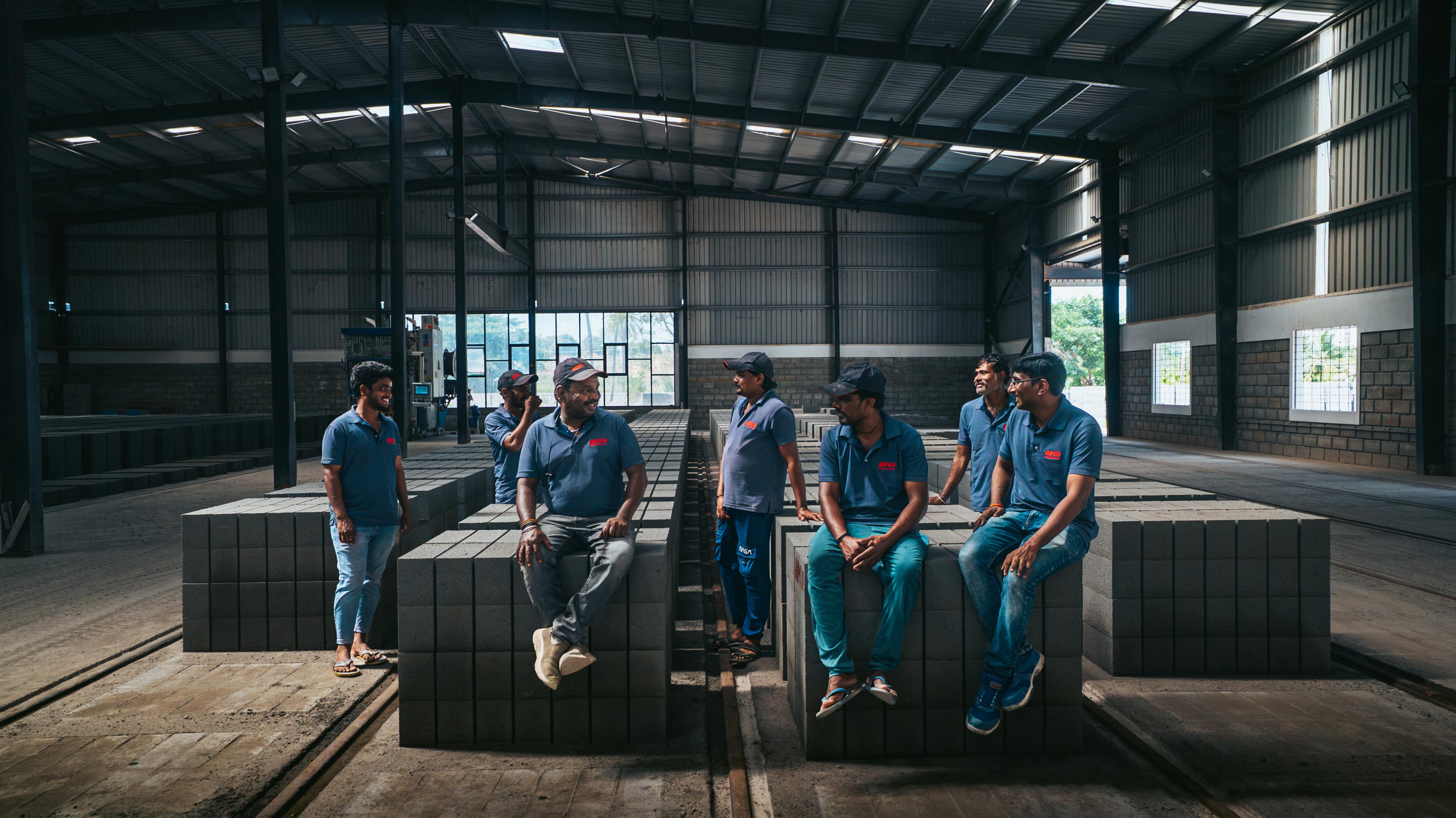 APCO factory team in branded polos sitting on stacks of concrete blocks inside the plant