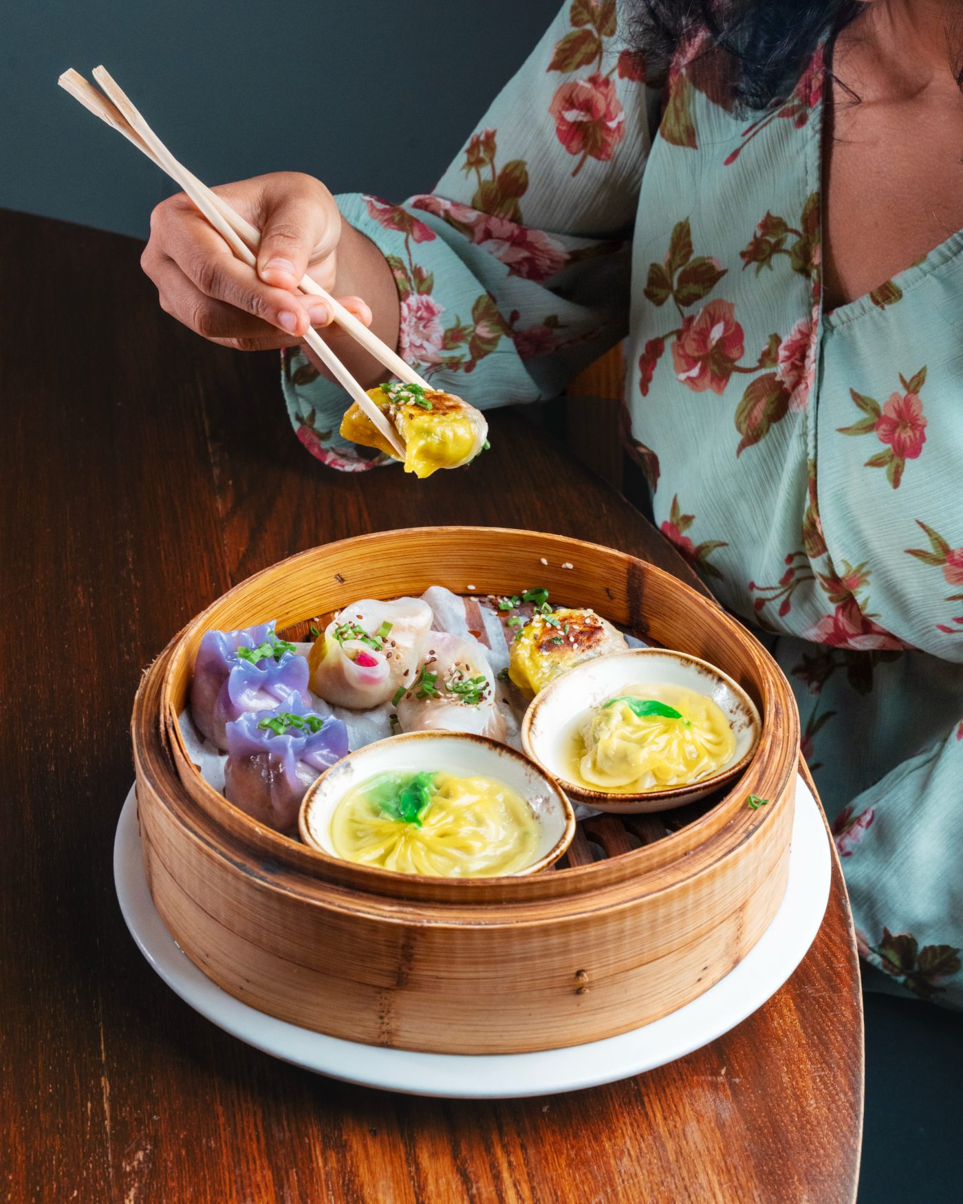 Diner lifting a dumpling with chopsticks over a bamboo steamer of colourful dim sum